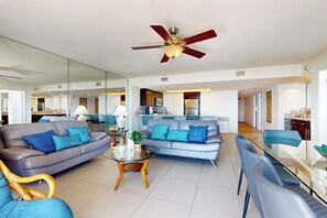 A modern living room with a glass coffee table, two sofas with blue cushions, a ceiling fan, and a kitchen in the background. - All-Suite Coastal Retreat: Suntide III - Unit 405 (South Padre Island)