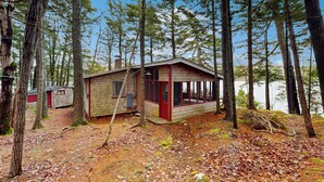 A cabin with a red door and windows, surrounded by tall trees and a forest floor covered with leaves. - Peaceful 2BR Lakefront Cabin w/ New Dock (Mount Desert)