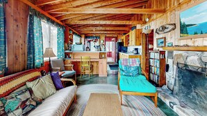 A cozy cabin interior with a wooden ceiling, a sofa, a reading chair, a fireplace, and a bookshelf. - Peaceful 2BR Lakefront Cabin w/ New Dock (Mount Desert)