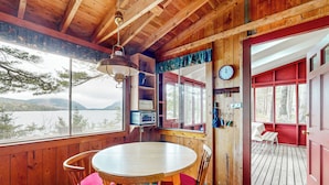 A wooden cabin with a round table and chairs, a clock, and a view of a lake through large windows. - Peaceful 2BR Lakefront Cabin w/ New Dock (Mount Desert)