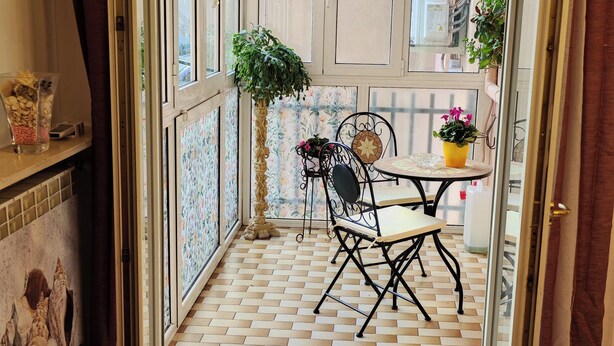 Un pequeño balcón cerrado con suelo de baldosas, una silla de metal, una mesita con planta en maceta y un panel decorativo en la pared.