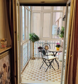 A small, enclosed balcony with a tiled floor, a metal chair, a small table with a potted plant, and a decorative wall panel.