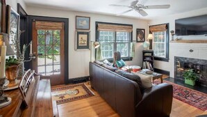 A living room with a dark leather sofa, wooden coffee table, and a fireplace. There are framed pictures on the walls and a ceiling fan. - Hideaway Hill lux downtown garden cottage & hottub (Waynesville)