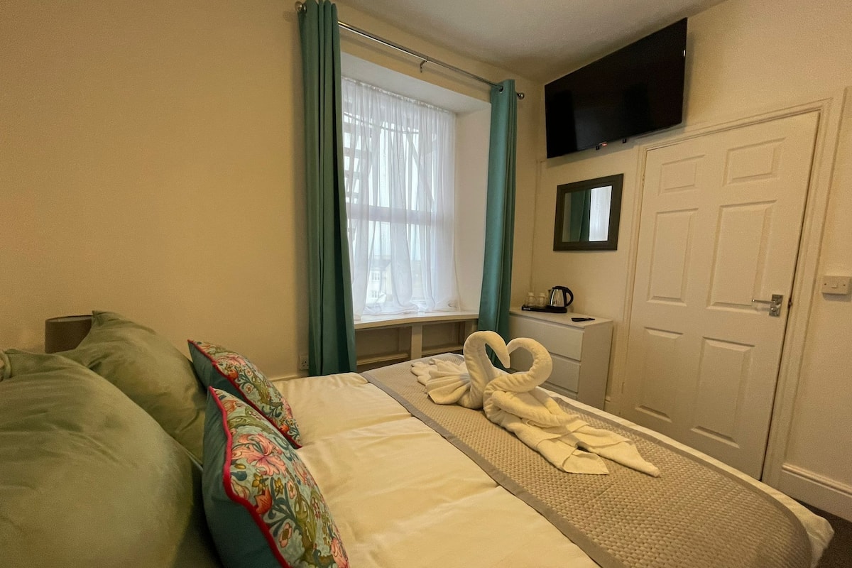Une chambre d’hôtel avec un lit, un bureau doté d’une bouilloire, un téléviseur fixé au mur et une porte.