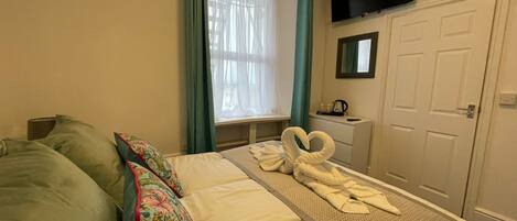 A hotel room with a bed, a desk with a kettle, a TV mounted on the wall, and a door.