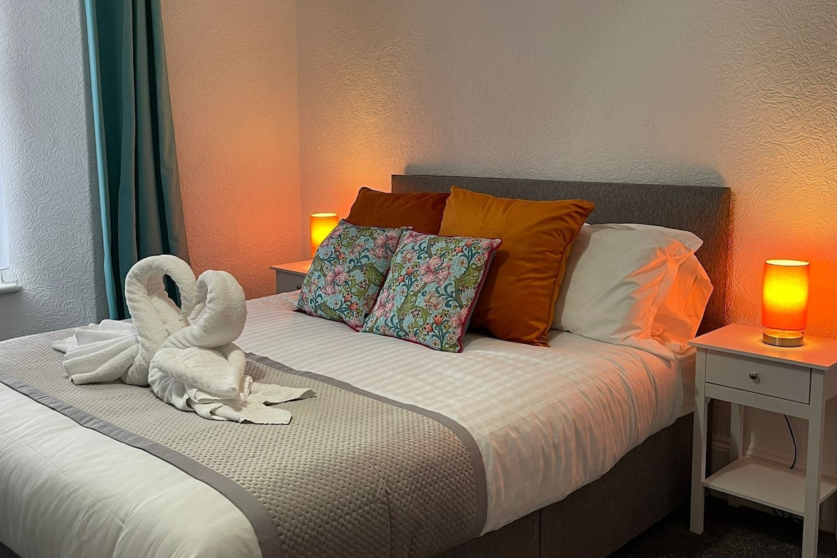 Un lit bien fait, recouvert d’une couette blanche, agrémenté de deux coussins décoratifs, d’une paire de serviettes blanches et d’une lampe de chevet posée sur une table de nuit blanche.