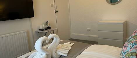 A hotel room with a bed, a TV mounted on the wall, a dresser, a mirror, and a towel arrangement on the bed.