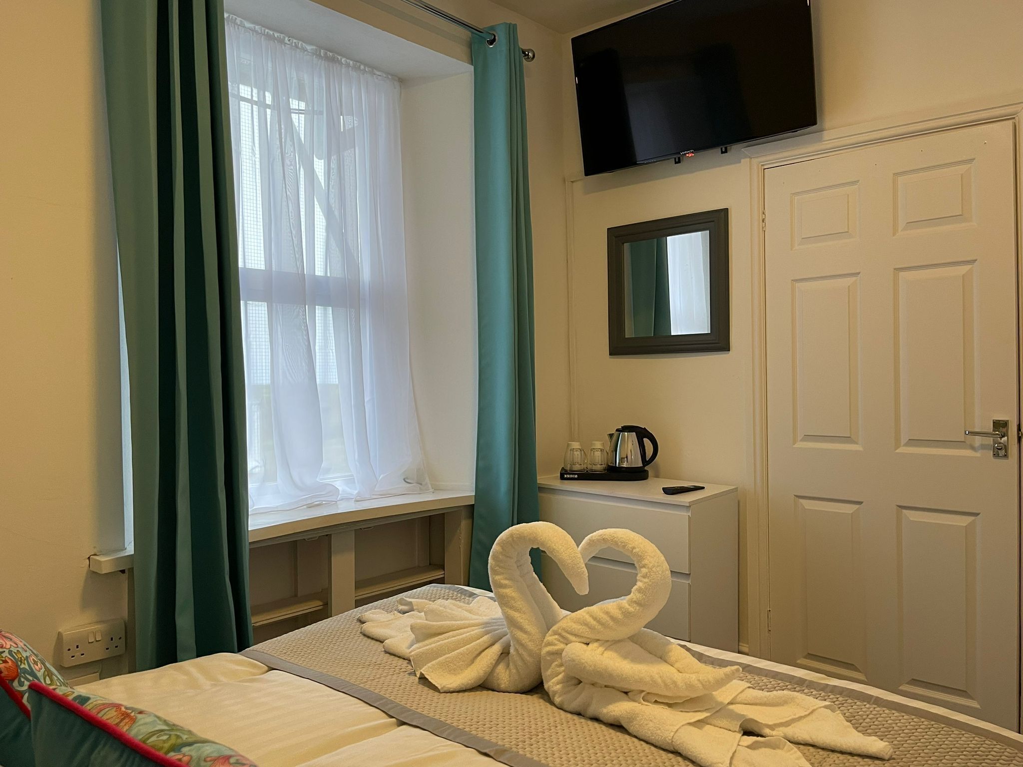 A hotel room with a bed, a window with sheer curtains, a flat-screen TV mounted above the bed, a small table with a kettle and cups, and a door with a mirror above it.
