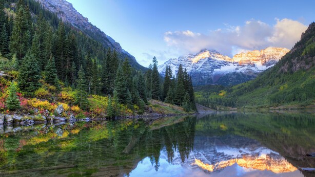Ein friedlicher Bergsee mit Spiegelungen von Bäumen und Bergen, umgeben von dichten Wäldern und felsigem Gelände.