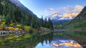 Un lago de montaña sereno que refleja árboles y montañas, rodeado de bosques densos y terreno rocoso.