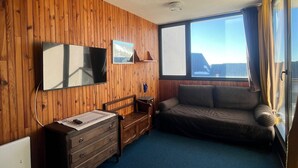 Ein Zimmer mit Holzwandverkleidung, einem großen Fenster mit Vorhängen, einem Sofa, einem Fernseher und einem Schminktisch.
