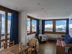 A cozy room with a wooden dining table, chairs, a bench, and a sofa. Large windows offer views of snow-covered mountains. - 2-room Apt, Plagne Centre, 5 ppl, slope view (La Plagne)