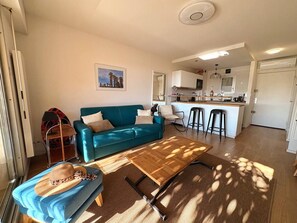 A living room with a teal sofa, a wooden coffee table, and a hat on a blue ottoman. A kitchen is visible in the background with a bar counter and stools. - T3 Apartment in Bormes-les-Mimosas - Near beaches and amenities (Bormes-les-Mimosas)
