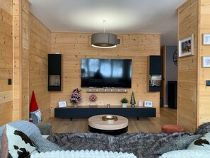 A modern living room with a large flat-screen TV mounted on the wall, wooden paneling on the walls, a round wooden coffee table, and a cozy seating area with a fur throw. - Modern apartment 8 pax, 3 bedrooms, parking, slope view (Pra-Loup)
