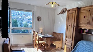 Een gezellige kamer met een houten eettafel, stoelen, een bankje, een blauwe bank, een houten kast en een raam met uitzicht op gebouwen en bomen.