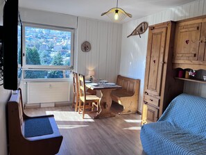 Une pièce chaleureuse avec une table à manger en bois, des chaises, un banc, un canapé bleu, un buffet en bois et une fenêtre donnant sur des bâtiments et des arbres.