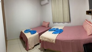 Une petite chambre d’hôtel avec deux lits simples, chacun recouvert d’une couvre-lit rose et agrémenté de serviettes bleues. Il y a une petite étagère en bois au-dessus du lit de droite, une fenêtre avec un rideau et un appareil de climatisation fixé au mur.