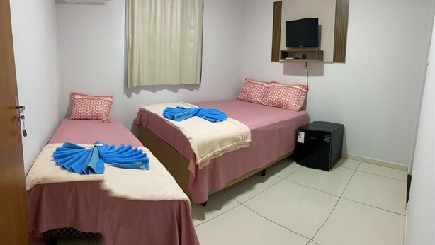 Une chambre avec deux lits, chacun recouvert d’une couvre-lit rose et agrémenté d’oreillers assortis. On y trouve un téléviseur fixé au mur, un petit meuble noir et une fenêtre avec un rideau blanc.