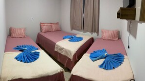Une chambre avec trois lits, chacun recouvert d’une couverture rose et agrémenté d’une serviette bleue. On y trouve une fenêtre avec des rideaux, une étagère en bois et une prise électrique au mur.