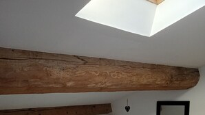 Um quarto com teto inclinado, uma viga de madeira, uma cama com colcha branca, um abajur e uma pequena mesa de cabeceira de madeira.