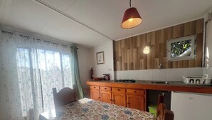 Eine kleine Küche mit Holzschränken, einem Gasherd und einem Spülbecken. Ein Esstisch mit blumigem Tischläufer und ein Stuhl stehen bereit.