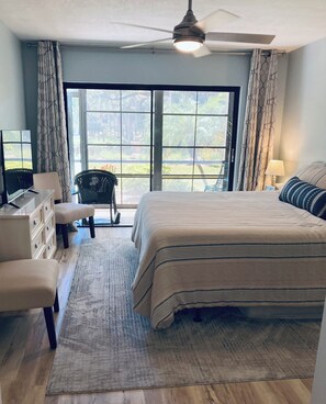 A bedroom with a large bed, a desk with a computer, a chair, and a view through a large window.