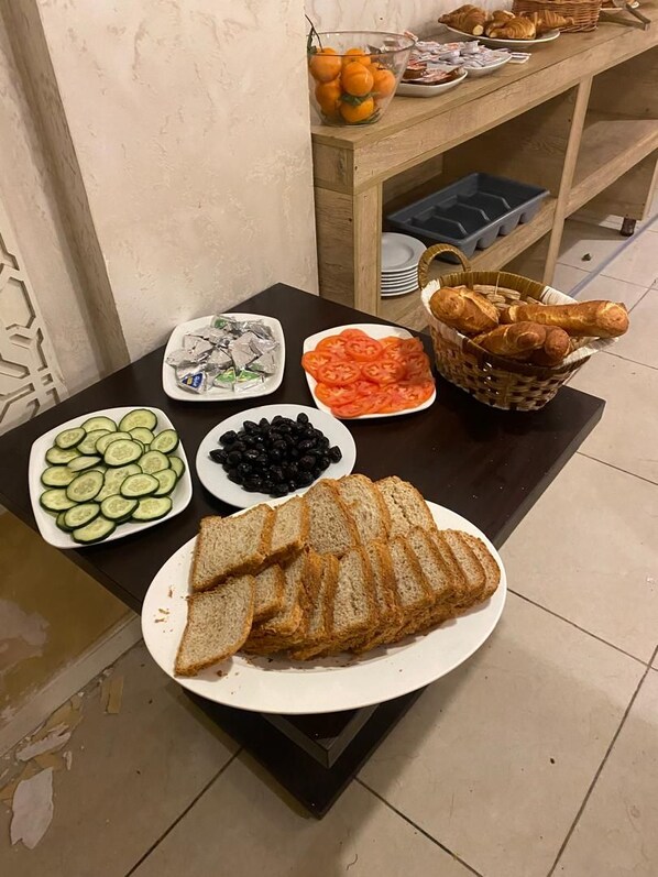 A buffet table with sliced cucumbers, black olives, tomatoes, bread, and a basket of croissants. - Hôtel kssar (Oran)