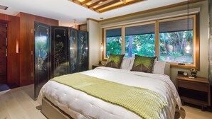Une chambre avec un grand lit, des poutres apparentes en bois au plafond, une fenêtre donnant sur la verdure et une table de chevet avec une horloge.