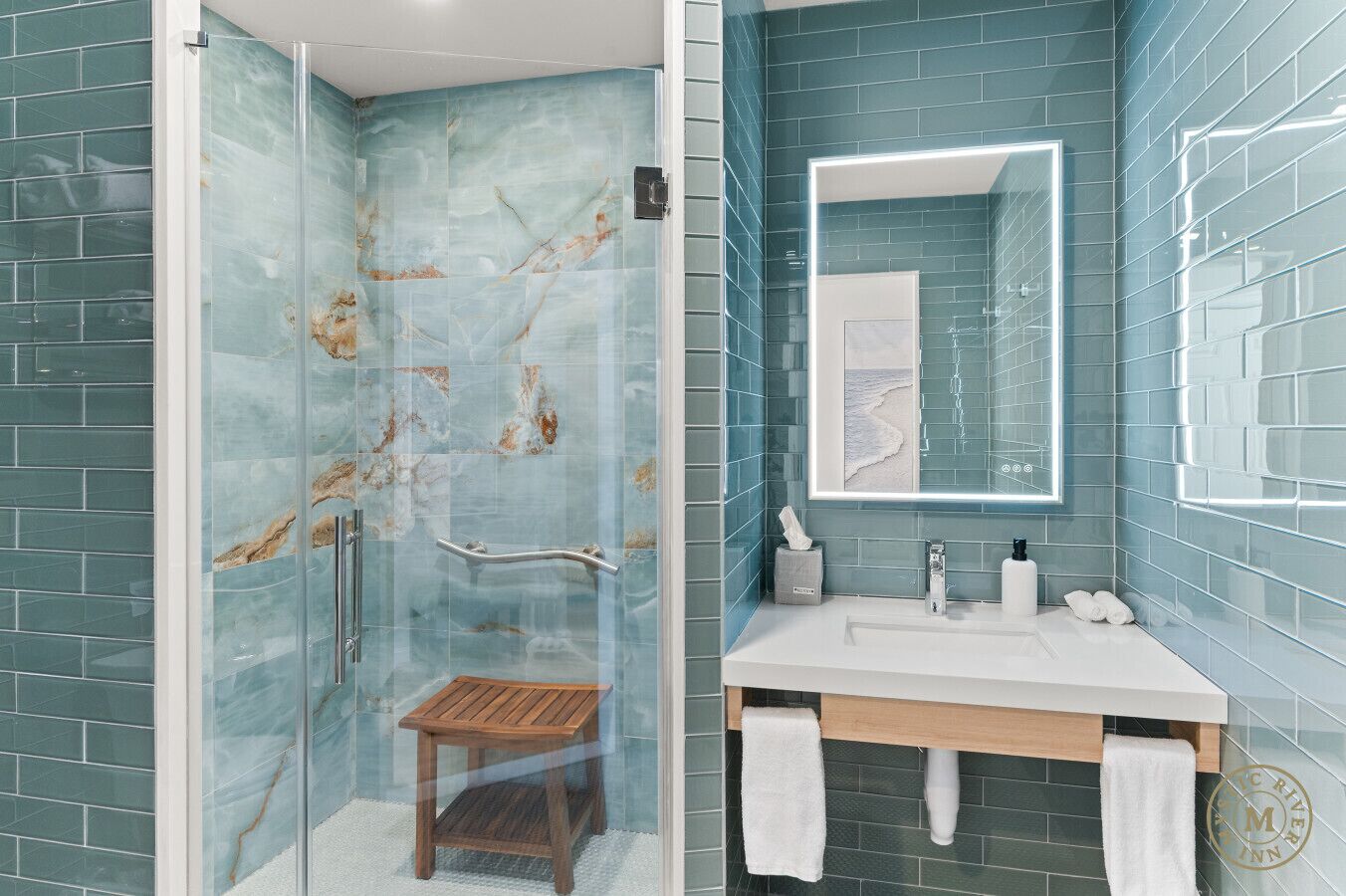 A bathroom with a glass shower enclosure, a wooden stool, a white sink, and a mirror with a light strip.