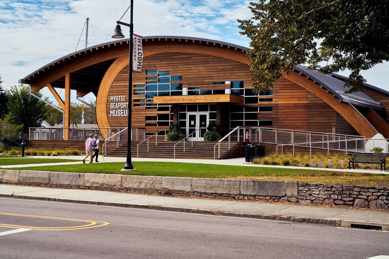 A modern building with a curved wooden facade, labeled as the Mystic Seaport Museum Thompson Exhibition Building.