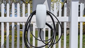 A Tesla charging station with a white fence and greenery in the background.