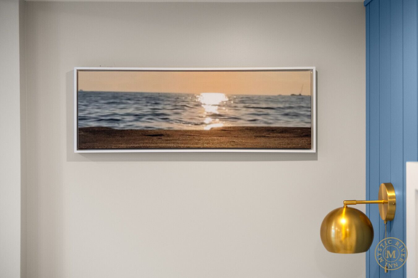 Ein gerahmtes Foto von einem Strand bei Sonnenuntergang an einer Wand, mit einer goldfarbenen Wandleuchte zur Rechten.