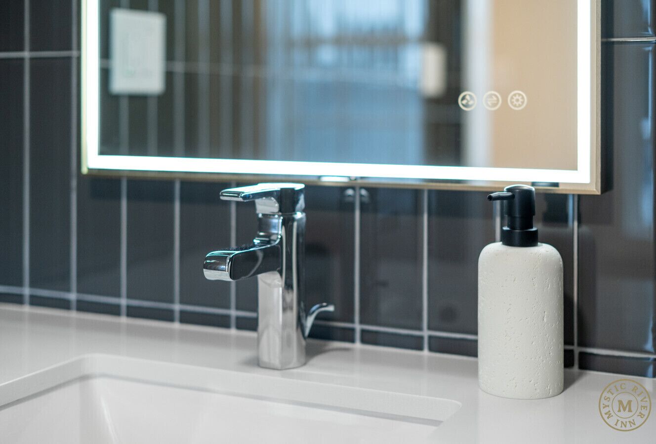 Een moderne badkamer met een chromen kraan, een witte zeepdispenser en een spiegel met verlichte rand.
