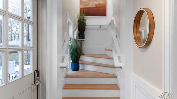 Una escalera con peldaños de madera y barandillas blancas, una planta en maceta en la plataforma y un espejo redondo en la pared.