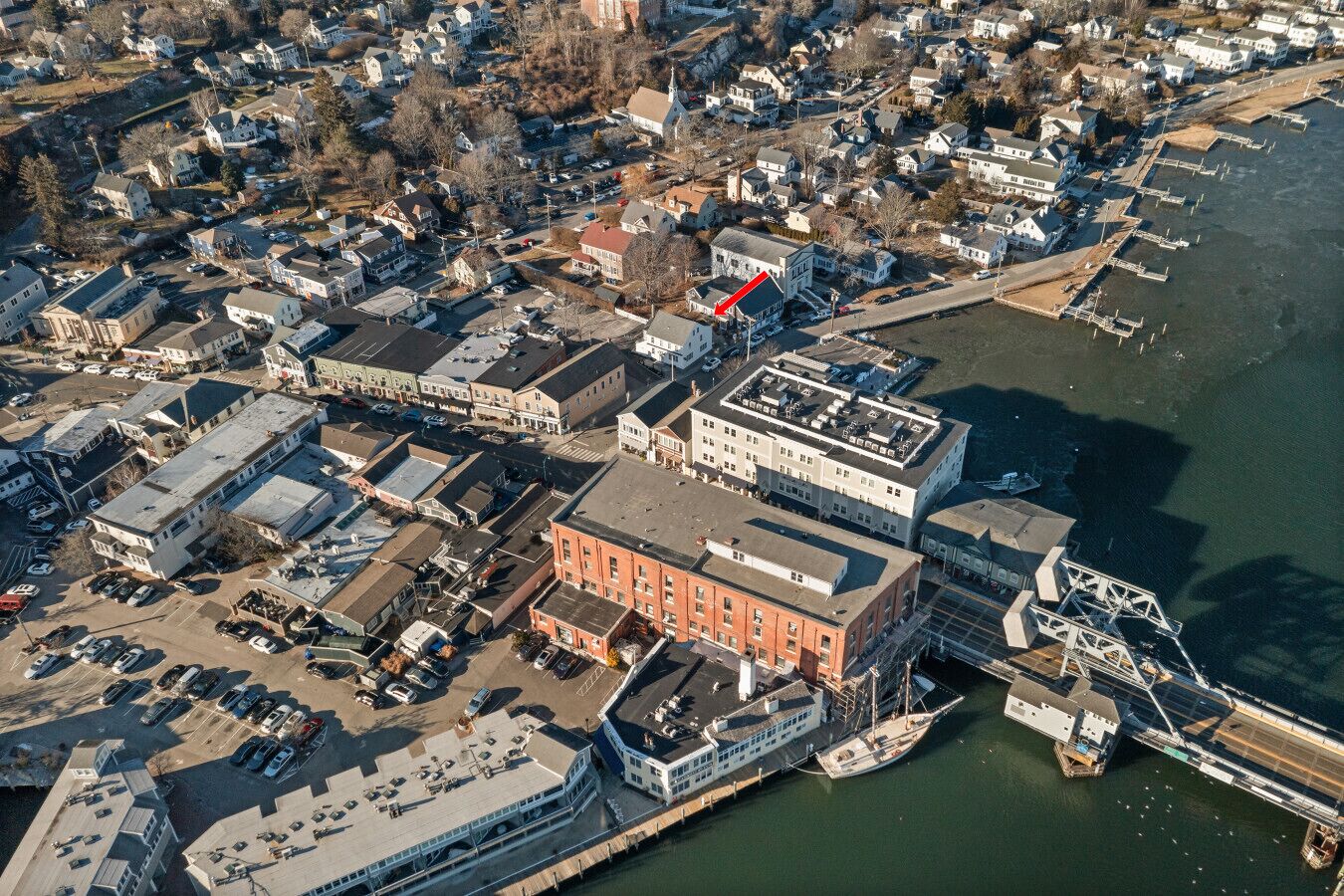 An aerial view of a waterfront area with a prominent red building, a marina with boats, and a bridge connecting to the land.