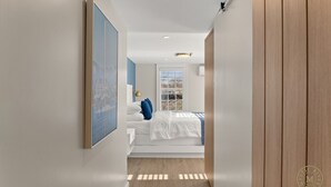 A narrow hallway leading to a bedroom with a bed, a window, and a framed picture on the wall. - The Mystic River Inn (Groton)