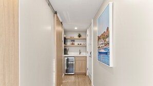 A narrow hallway leading to a compact kitchen with a sink, cabinets, and a refrigerator. A framed picture of a coastal scene hangs on the wall. - The Mystic River Inn (Groton)