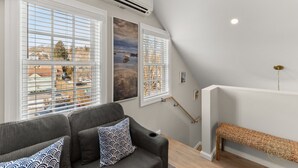 Una sala de estar moderna con un sofá gris, cojines estampados, ventanas amplias con persianas, un equipo de aire acondicionado y un banco de madera.