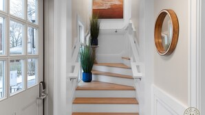 Una escalera con peldaños de madera y barandillas blancas, una planta en maceta en la plataforma y un espejo redondo en la pared.