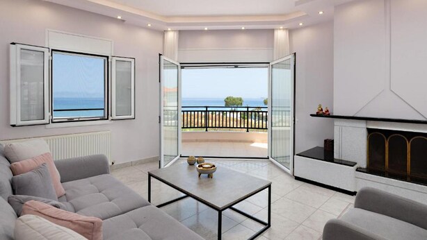 Ein modernes Wohnzimmer mit einer Couch, einem Couchtisch und einem Kamin. Ein großes Glasfenster führt auf einen Balkon mit Meerblick.
