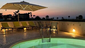 Ein Pool im Freien mit Liegestühlen und Sonnenschirm, umgeben von einer niedrigen Mauer und Palmen im Hintergrund.