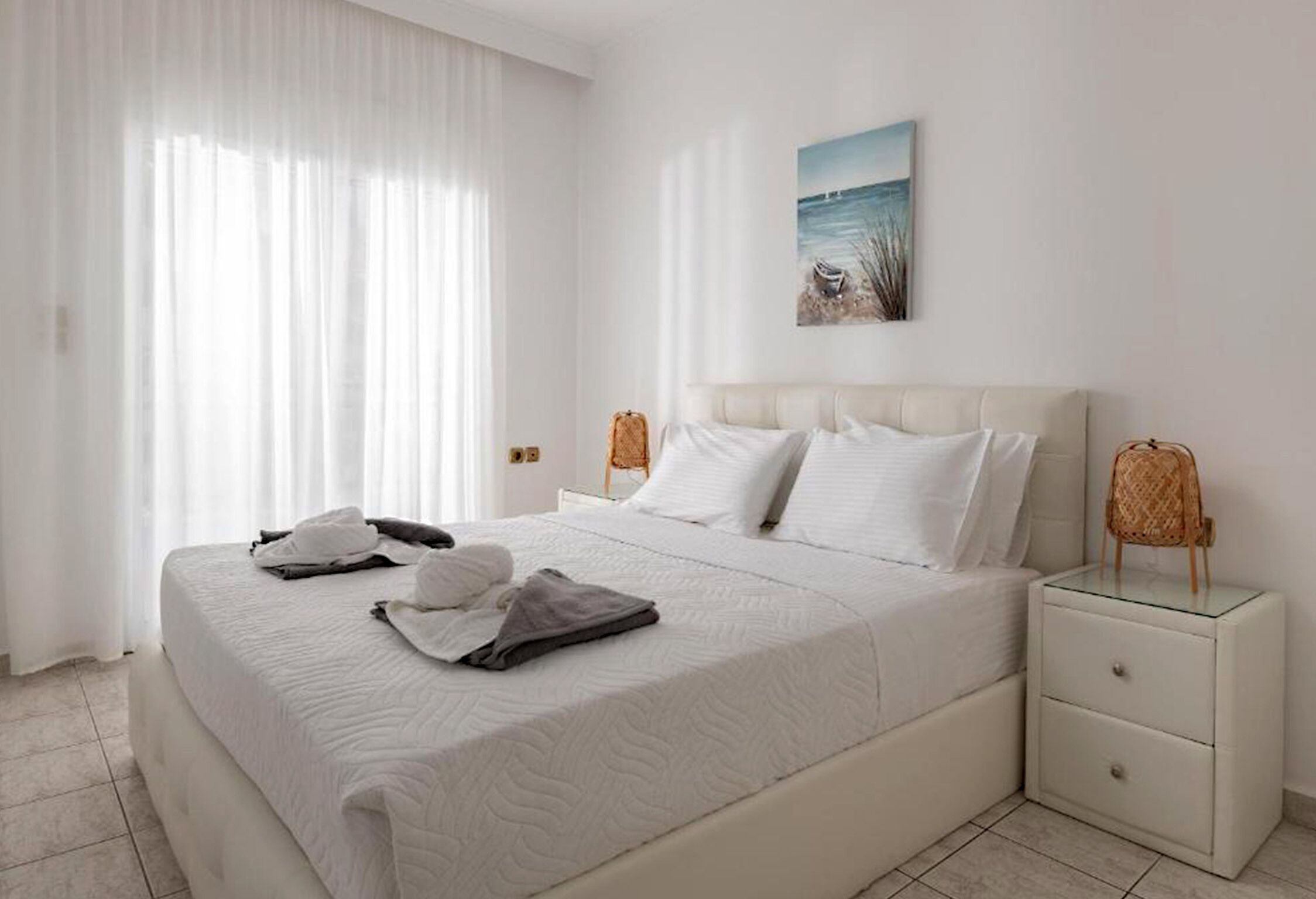 A neatly made bed with white linens and pillows, a bedside table with a lamp, and a painting of a beach scene on the wall.