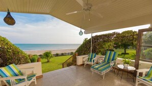 Un'area esterna riparata con vista sulla spiaggia, caratterizzata da sedie bianche con cuscini a righe, un tavolino con una pianta e una ventola a soffitto.