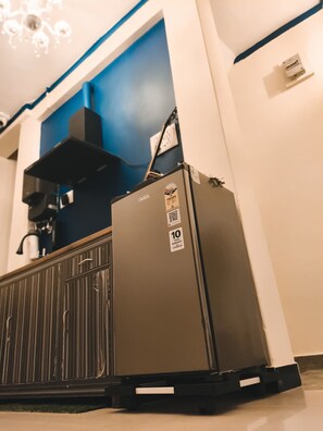 A compact refrigerator with a warranty sticker, placed in a room with a blue accent wall and a radiator. - Veena Vihar (Full House) (Jodhpur)