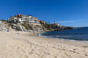 Ein Strand mit einem Hotel auf einer felsigen Klippe, blauem Himmel und ruhigem Meer.