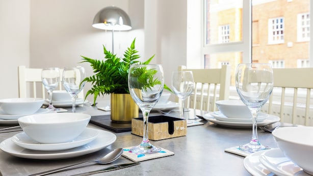 A dining table set with white plates, bowls, and clear wine glasses, with a potted plant and a lamp in the background. - A Nice Apartment in Central London, Great For Sightseeing (London)