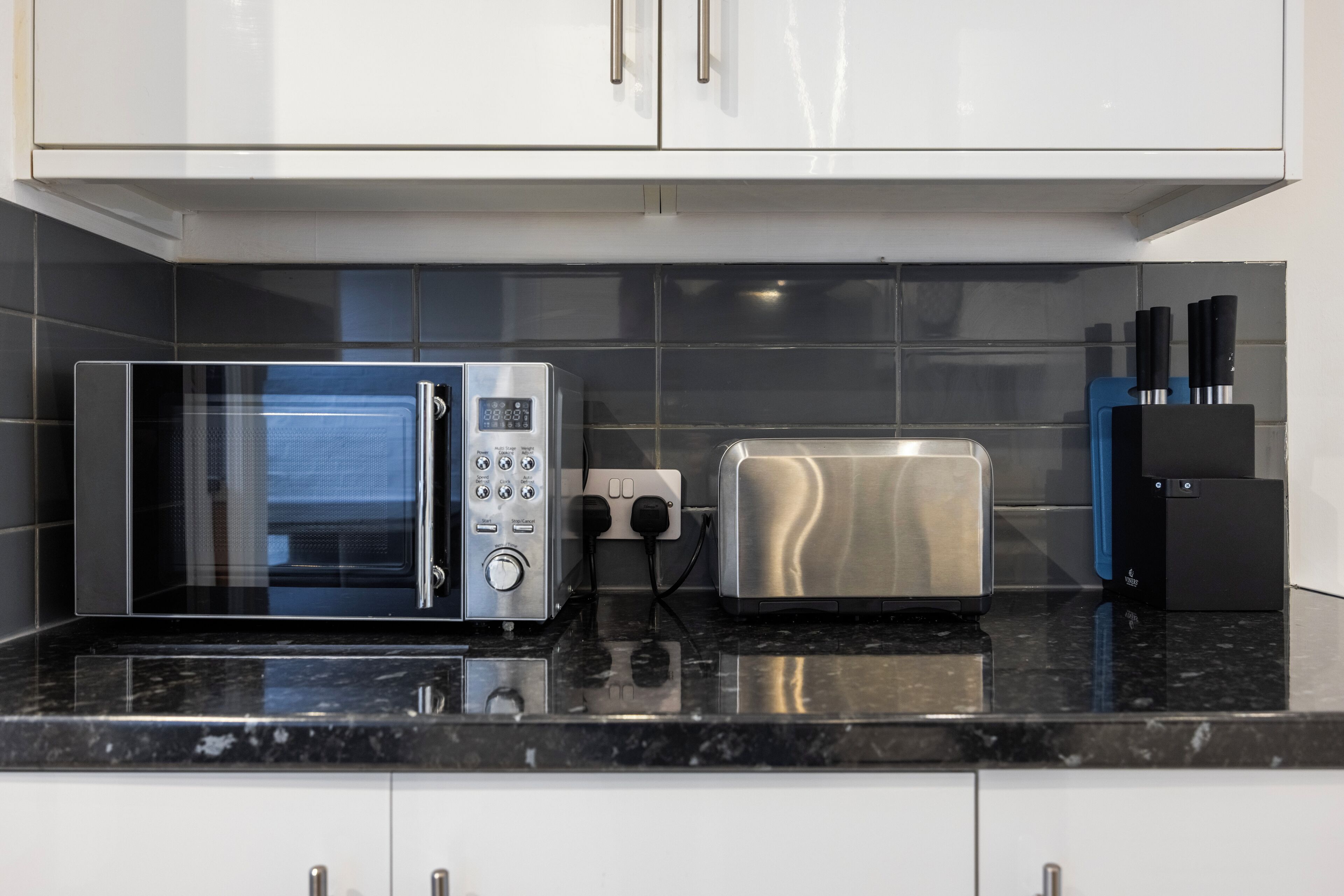 Eine Küche mit Mikrowelle, Toaster und einem schwarzen Gerät, möglicherweise eine Kaffeemaschine oder Speisewärmer, auf einer schwarzen Arbeitsplatte.