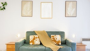 Een kamer met een blauwgroene bank, twee kussens met een bladmotief, een beige plaid en drie bladmotieven in lijsten aan de muur.