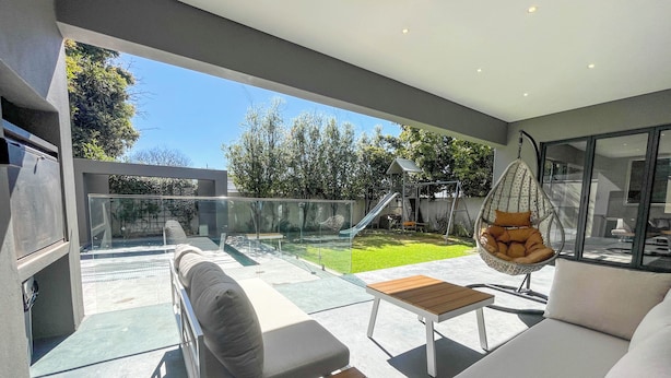 A modern outdoor living space with a glass balustrade, a hanging chair, and a play area visible through the glass.