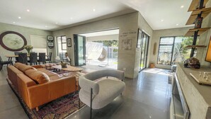 A modern living room with a brown leather sofa, a grey armchair, and a coffee table. There is a dining area with a table and chairs, and a large window providing natural light.